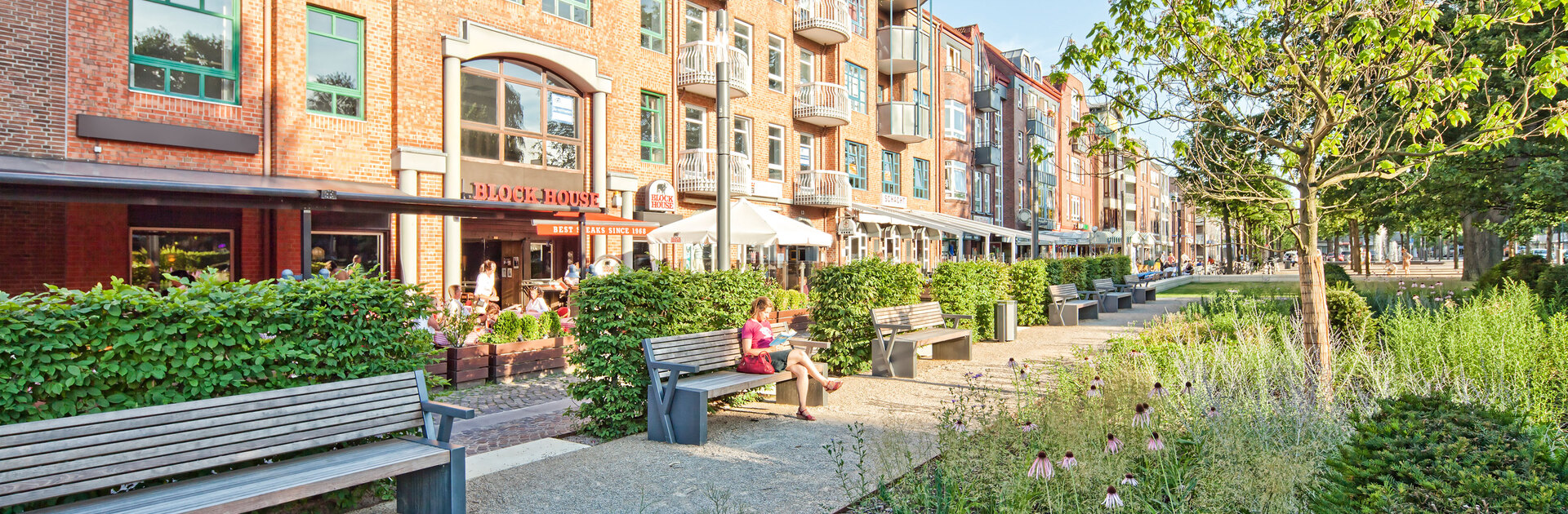 Blick von einem grün gesäumten Fußweg auf das BLOCK HOUSE Restaurant in Ahrensburg. Im Vordergrund stehen mehrere Bänke, auf einer sitzt eine Frau. Vor dem Restaurant ist eine Terrasse, auf der mehrere Gäste sitzen. 