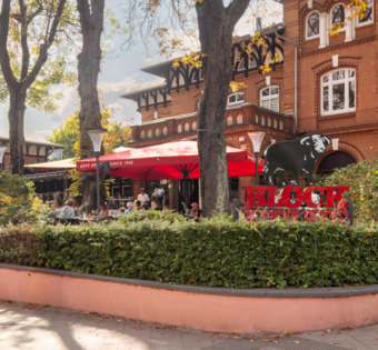 BLOCK HOUSE Hamburg Othmarschen – Steakhouse mit Terrasse BLOCK HOUSE Hamburg Othmarschen mit Terrasse, roten Sonnenschirmen und Gästen vor dem Steakhouse im grünen Umfeld
