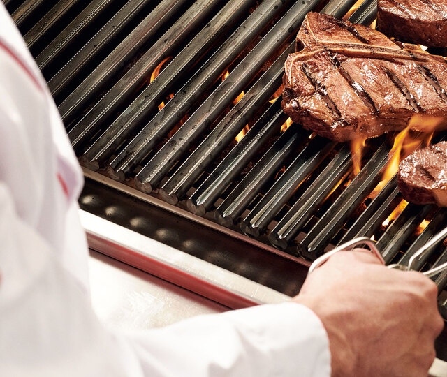 Blick über die Schulter eines Grillers, der eine Steakzange in der Hand hält und gerade ein Steak auf einem flammenden Grill wendet.