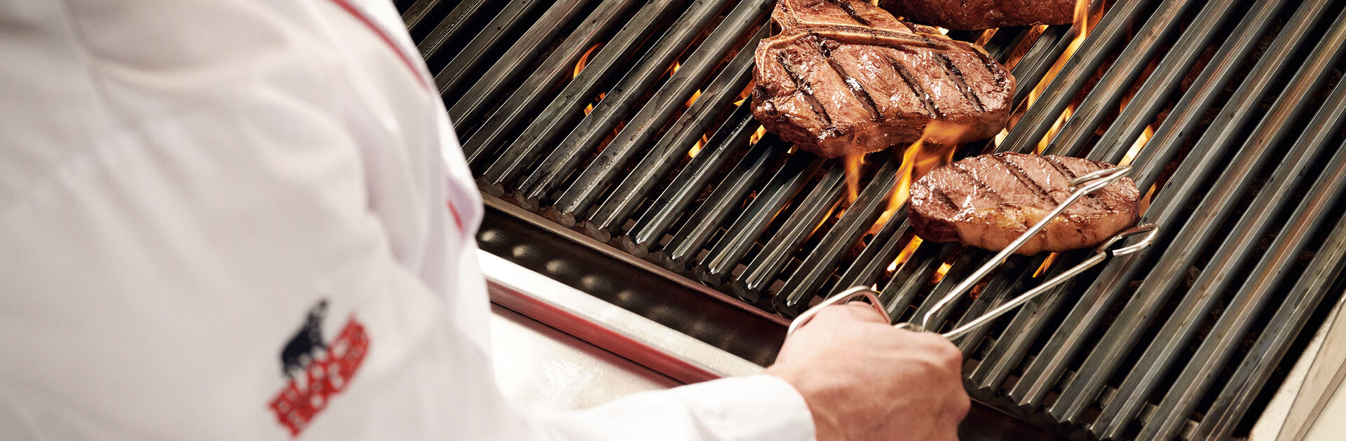 Blick über die Schulter eines Grillers, der eine Steakzange in der Hand hält und gerade ein Steak auf einem flammenden Grill wendet.