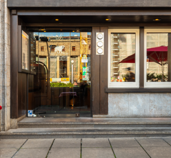 BLOCK HOUSE Stuttgart Arnulf-Klett-Platz – Steakhouse mit Terrasse Eingang des BLOCK HOUSE Stuttgart Arnulf-Klett-Platz mit Menütafel vor dem Steakhouse und Gästen an Tischen mit rot-weiß karierten Tischdecken
