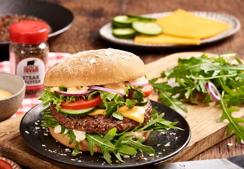 BLOCK HOUSE Burger mit Fleischpatty, Rucola, Tomate, Salat , Zwiebeln und Sauce auf einem Holzbrett mit verwendeten Zutaten im Hintergrund