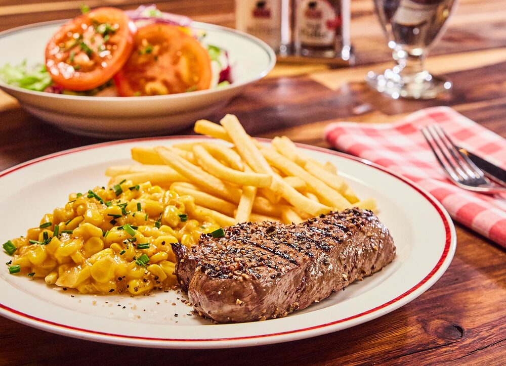 Huftsteak mit Pommes und Maisgemüse auf einem Teller serviert, dazu ein Salat 