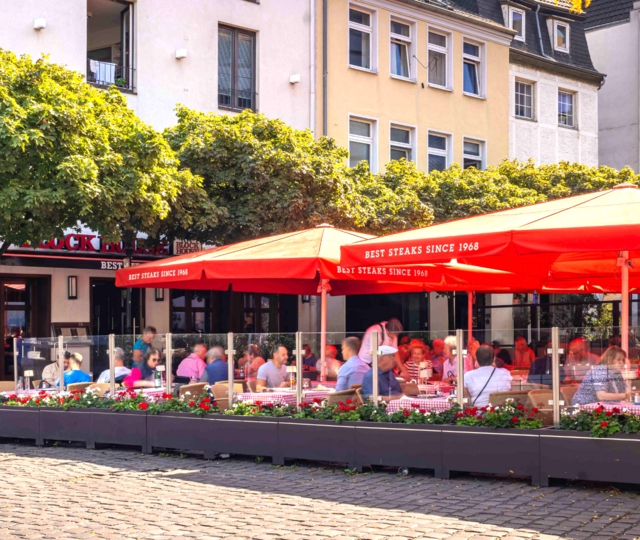Außenansicht des BLOCK HOUSE Düsseldorf am Burgplatz mit großer Terrasse, roten Sonnenschirmen und Gästen.