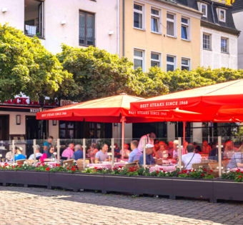 Außenansicht des BLOCK HOUSE Düsseldorf am Burgplatz mit großer Terrasse, roten Sonnenschirmen und Gästen.