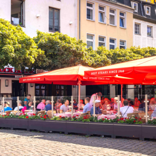 Außenansicht des BLOCK HOUSE Düsseldorf am Burgplatz mit großer Terrasse, roten Sonnenschirmen und Gästen.