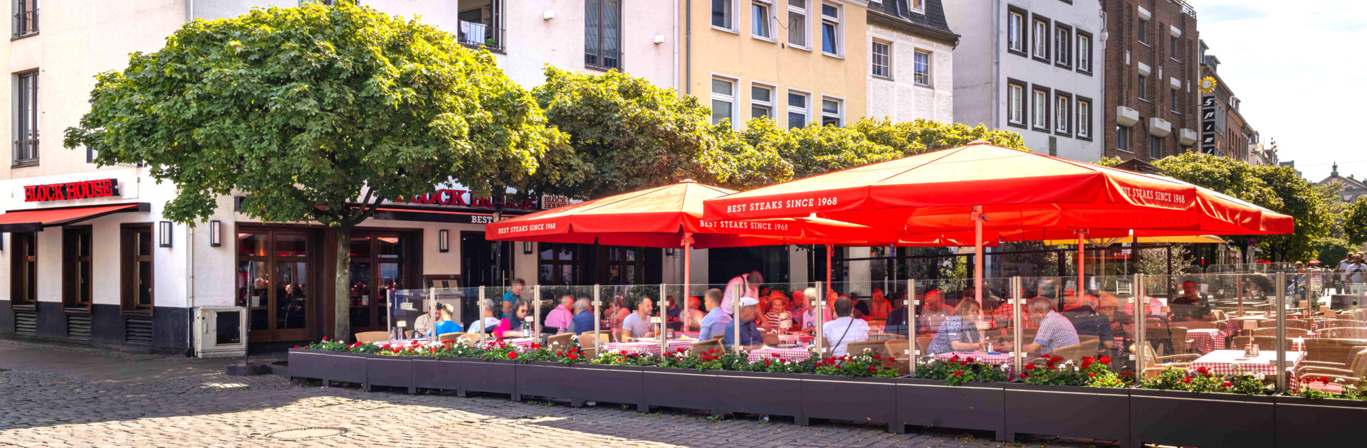 Außenansicht des BLOCK HOUSE Düsseldorf am Burgplatz mit großer Terrasse, roten Sonnenschirmen und Gästen.