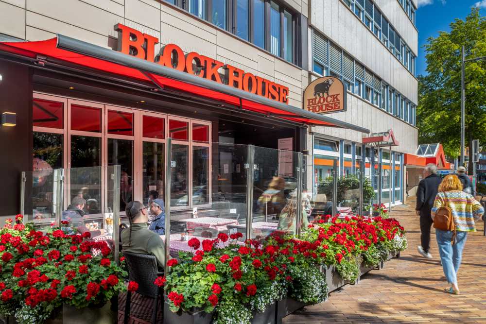 BLOCK HOUSE Hamburg Harburg: Gäste genießen Steaks auf der Außenterrasse mit roter Markise und Geranien bei strahlendem Sonnenschein.