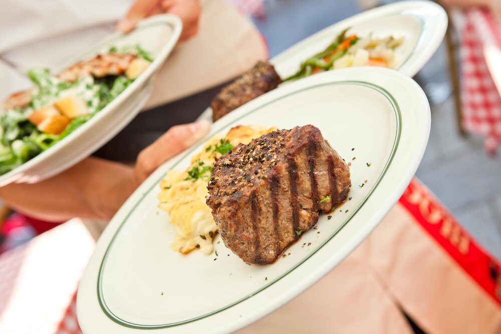 Verschiedene Gerichte werden von einer Kellnerin im BLOCK HOUSE serviert. Im Vordergrund ein Steak auf einem Teller. 