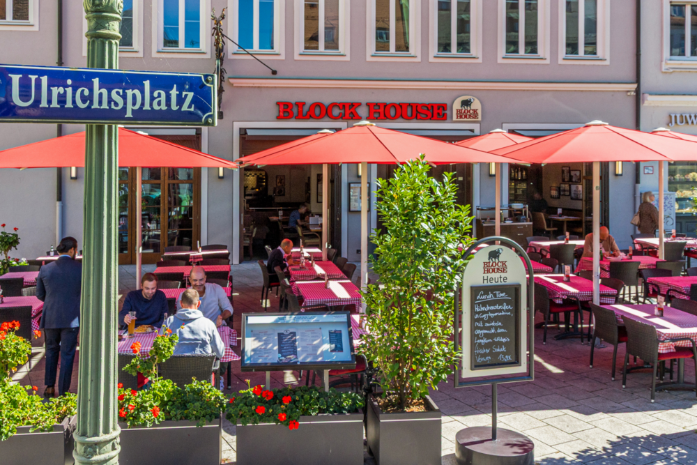 Außenansicht des BLOCK HOUSE Augsburg am Ulrichsplatz mit Terrasse und roten Sonnenschirmen.