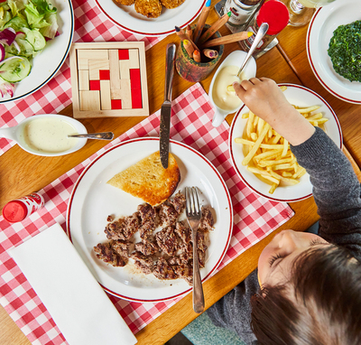 Kind sitzt am Tisch und isst. Auf dem Tisch liegen Spielzeug und Malstifte 