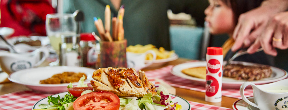 Ein frischer Salat mit gegrilltem Hähnchen im Block House, während im Hintergrund ein Steak serviert wird und ein Kind unscharf zu erkennen ist.