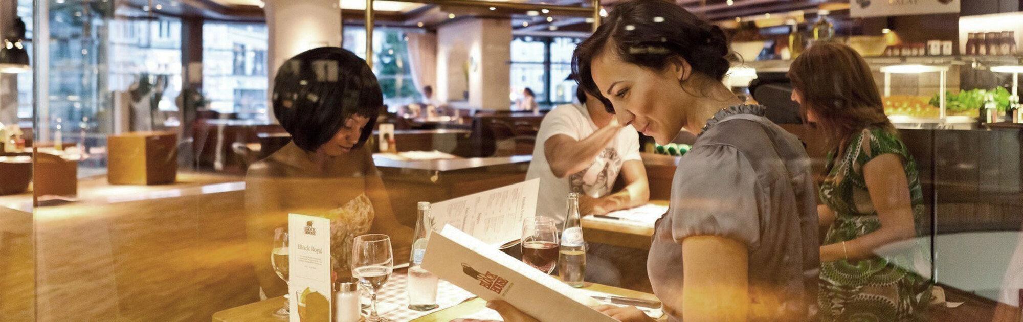 Eine Frau sitzt im BLOCK HOUSE Restaurant und liest aufmerksam die Speisekarte. Auf dem Tisch liegen Besteck, ein Glas Wasser, ein Menüaufsteller und ein kariertes Platzset.