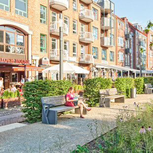 Blick von einem grün gesäumten Fußweg auf das BLOCK HOUSE Restaurant in Ahrensburg. Im Vordergrund stehen mehrere Bänke, auf einer sitzt eine Frau. Vor dem Restaurant ist eine Terrasse, auf der mehrere Gäste sitzen. 