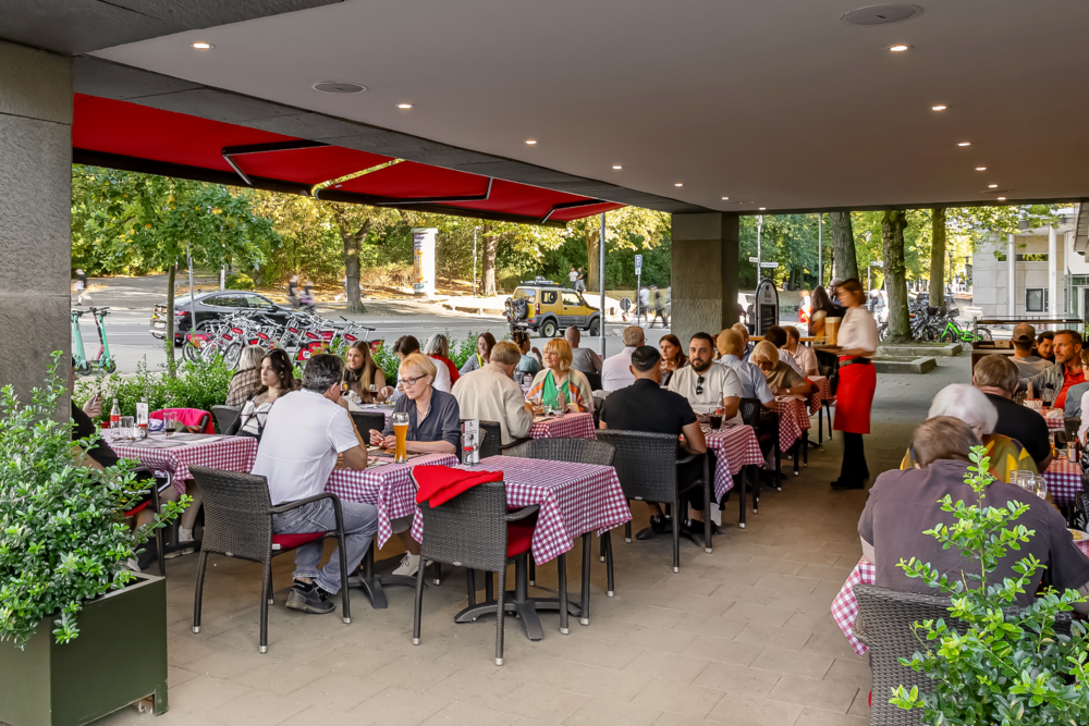 Gäste sitzen auf der Terrasse des BLOCK HOUSE Braunschweig an Tischen mit rot-weiß karierten Tischdecken, Servicekraft bringt Getränke