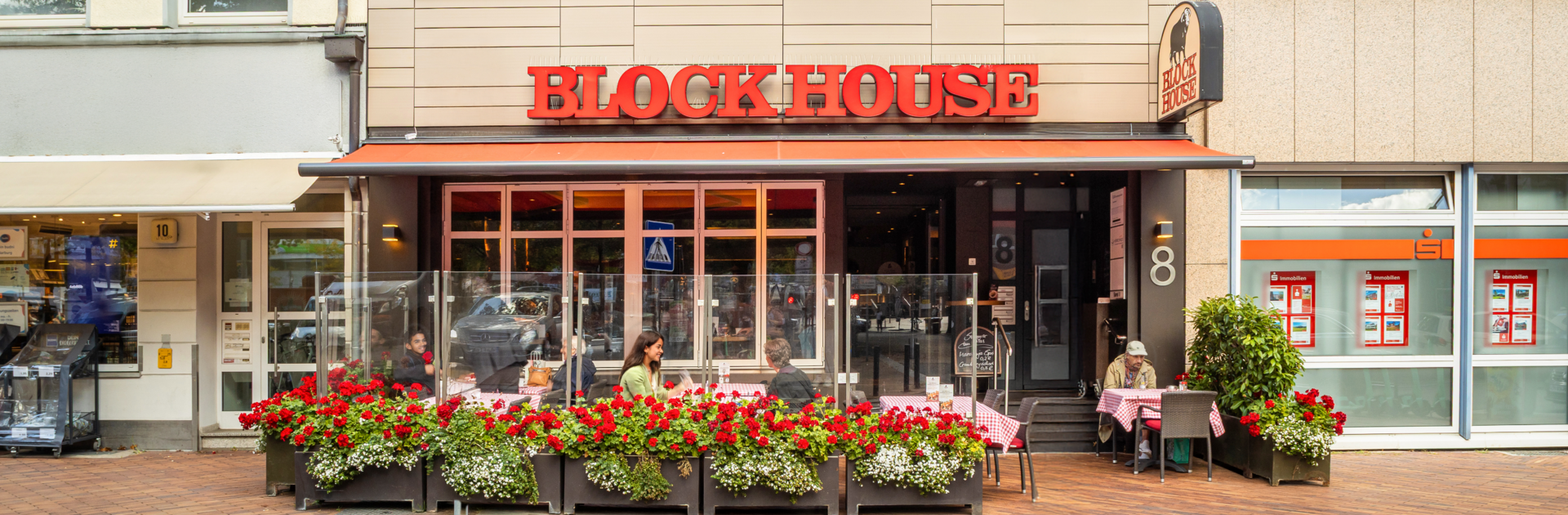 Eingangsbereich des Steak-Restaurants BLOCK HOUSE Hamburg Harburg mit Außenterrasse, einer roten Markise und blühenden Geranien.