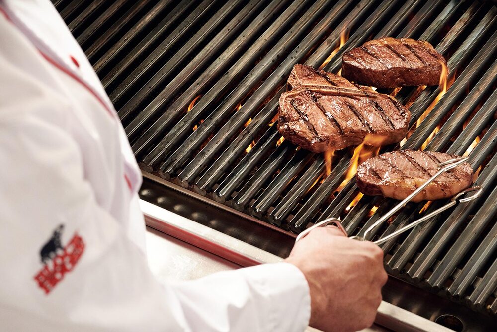 Blick über die Schulter eines Grillers, der eine Steakzange in der Hand hält und gerade ein Steak auf einem flammenden Grill wendet.