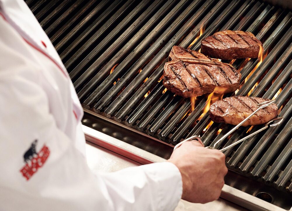 Blick über die Schulter eines Grillers, der eine Steakzange in der Hand hält und gerade ein Steak auf einem flammenden Grill wendet.