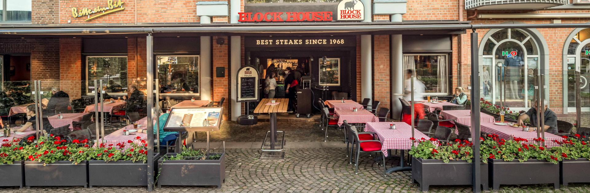 Terrasse des BLOCK HOUSE Ahrensburg mit Gästen an Tischen mit rot-weiß karierten Tischdecken vor dem Steakhouse