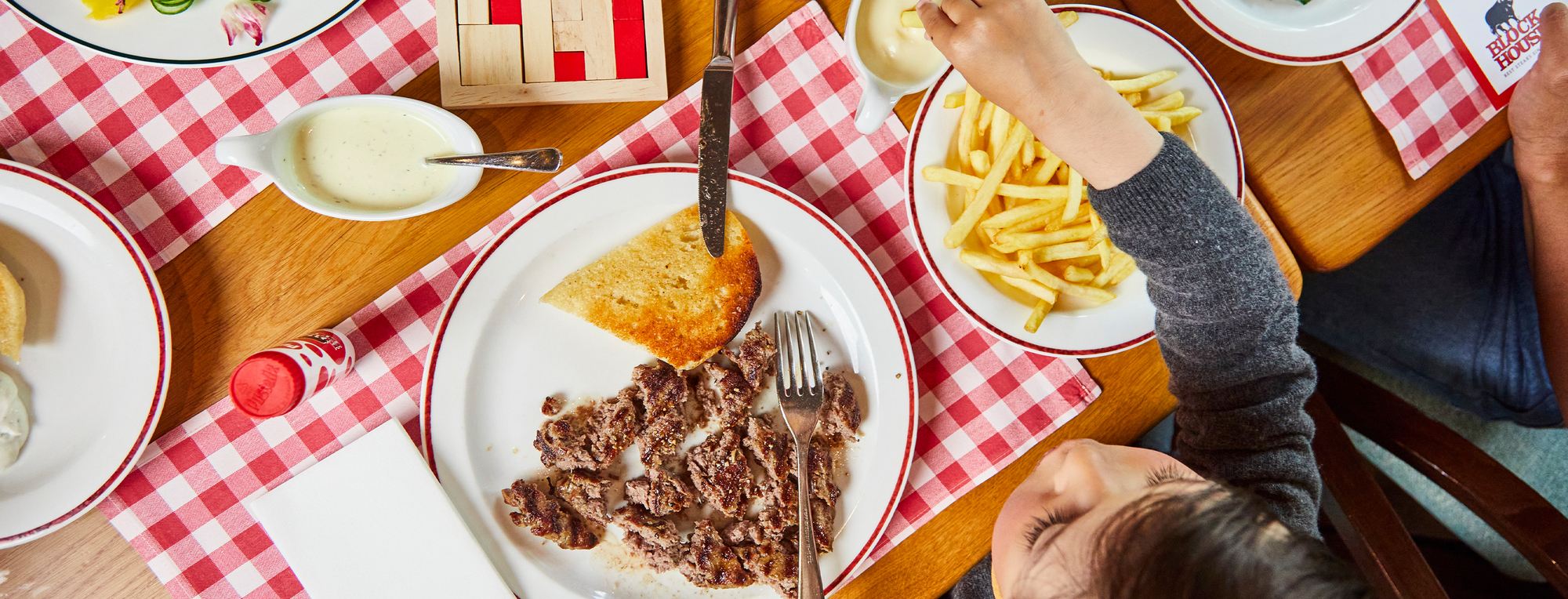 Ein Kind greift nach Pommes auf einem Teller, während daneben ein teilweise gegessenes Steak mit Brot liegt. Der Tisch im BLOCK HOUSE Restaurant ist mit einer rot-weiß karierten Tischdecke gedeckt und mit verschiedenen Speisen, Saucen und Besteck versehen.