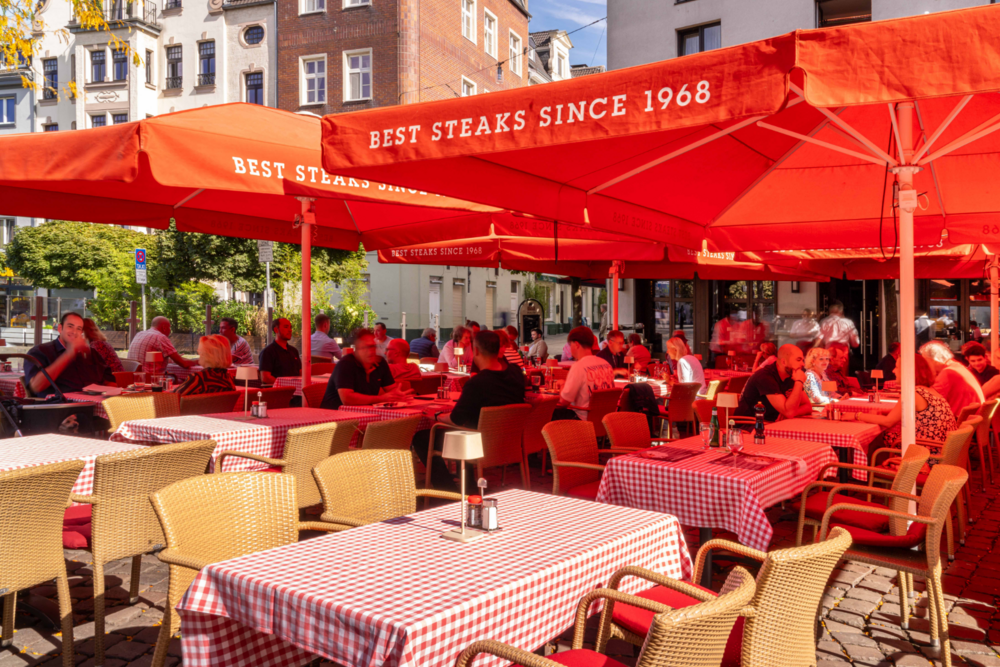 Terrasse des BLOCK HOUSE Düsseldorf am Burgplatz mit roten Sonnenschirmen und Gästen im Außenbereich.