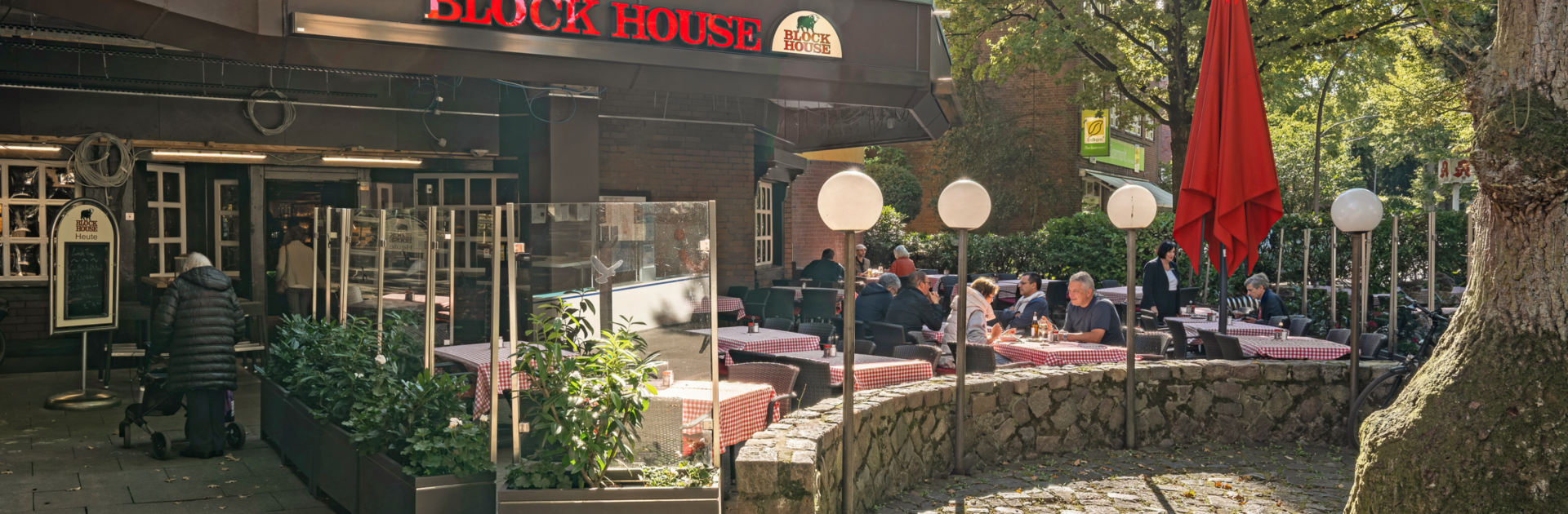 BLOCK HOUSE Hamburg Volksdorf mit Gästen vor dem Steakhouse und Sitzplätzen auf der Terrasse unter großen Bäumen