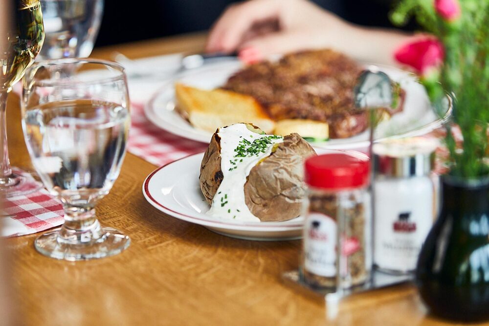 Blick auf einen gedeckten Tisch im BLOCK HOUSE: Ein Glas Wasser, eine Baked Potato mit Sour Cream, Gewürze und im Hintergrund ein Steakteller
