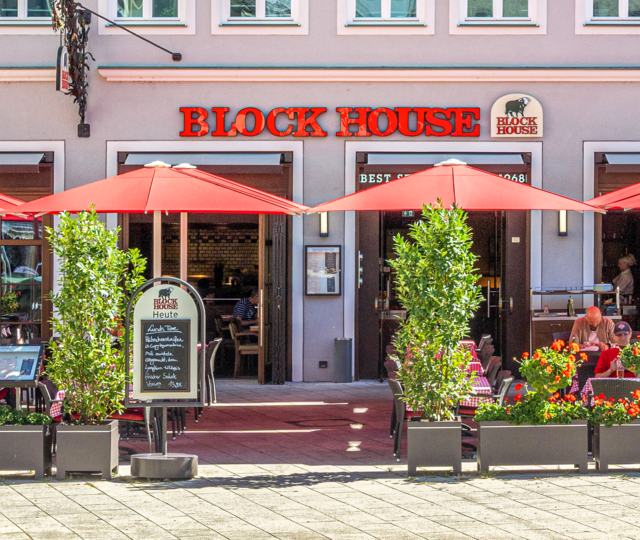 Außenansicht des BLOCK HOUSE Restaurants in Augsburg: eine große, hölzerne Fensterfront, davor viele Sitzgelegenheiten auf der Terrasse, die sich direkt auf dem Ulrichsplatz befindet.