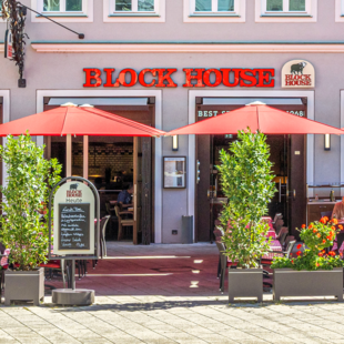 BLOCK HOUSE Restaurant Augsburg Außenansicht des BLOCK HOUSE Restaurants in Augsburg: eine große, hölzerne Fensterfront, davor viele Sitzgelegenheiten auf der Terrasse, die sich direkt auf dem Ulrichsplatz befindet.