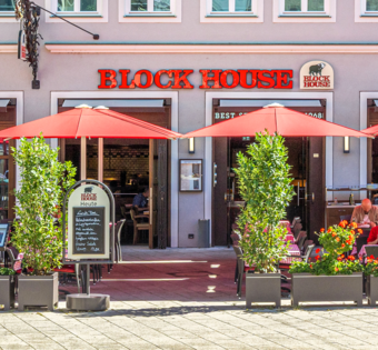 Außenansicht des BLOCK HOUSE Restaurants in Augsburg: eine große, hölzerne Fensterfront, davor viele Sitzgelegenheiten auf der Terrasse, die sich direkt auf dem Ulrichsplatz befindet.