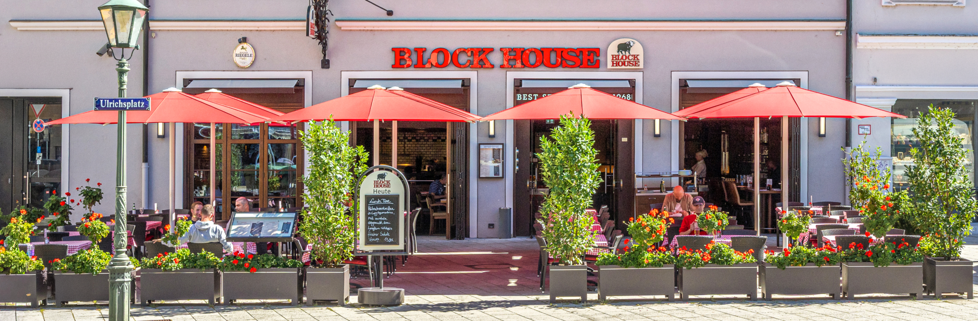 Außenansicht des BLOCK HOUSE Restaurants in Augsburg: eine große, hölzerne Fensterfront, davor viele Sitzgelegenheiten auf der Terrasse, die sich direkt auf dem Ulrichsplatz befindet.
