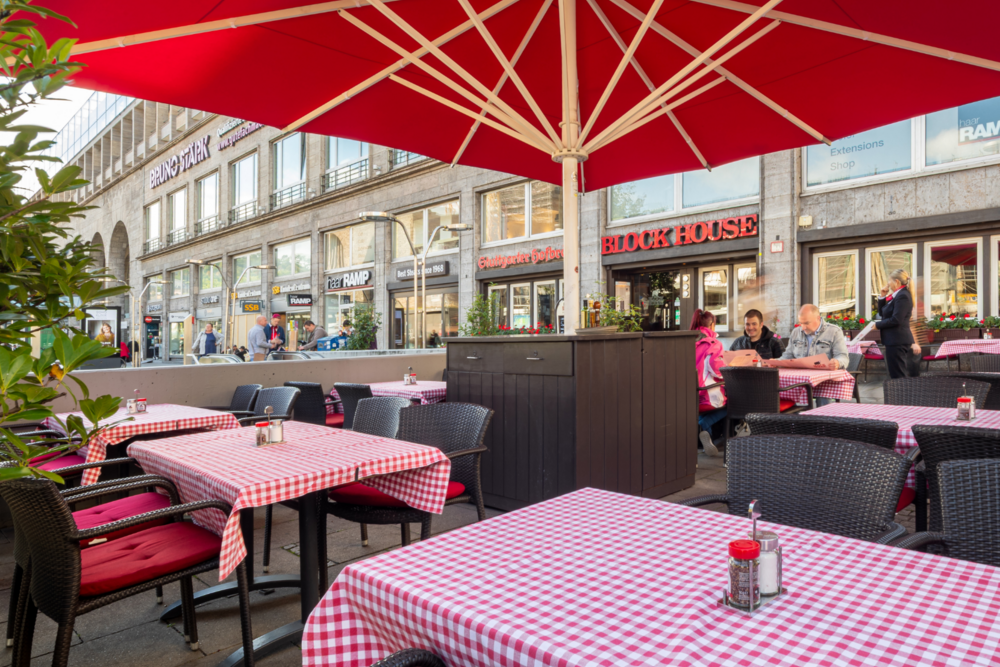 Terrasse des BLOCK HOUSE Stuttgart Arnulf-Klett-Platz mit Tischen mit rot-weiß karierten Tischdecken unter rotem Sonnenschirm