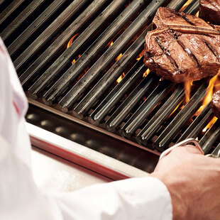 Blick über die Schulter eines Grillers, der eine Steakzange in der Hand hält und gerade ein Steak auf einem flammenden Grill wendet.