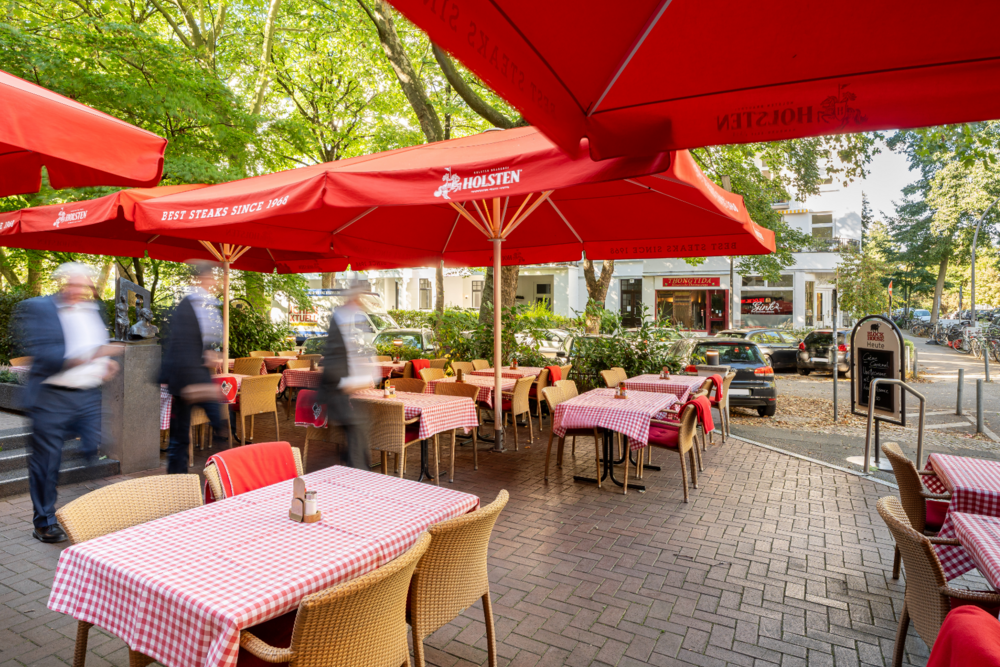 Außenansicht der Terrasse vom BLOCK HOUSE Hamburg-Winterhude mit gedeckten Tischen, roten Schirmen und großen Bäumen