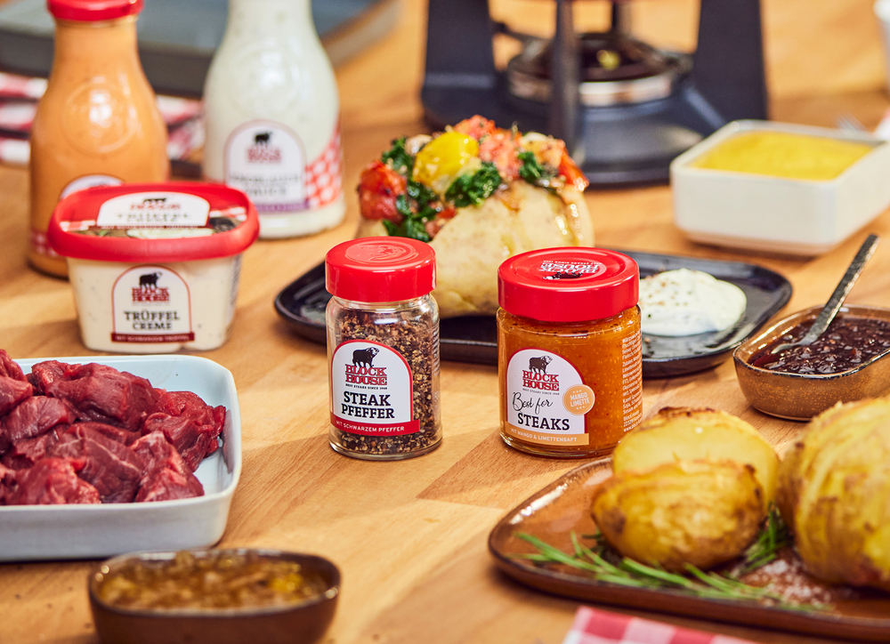 BLOCK HOUSE Fleisch, Saucen und Gewürze stehen mit zubereiteten Hasselback und Garden Potatoes auf einem Holztisch. Im Hintergrund ist ein Fondue zu sehen. 