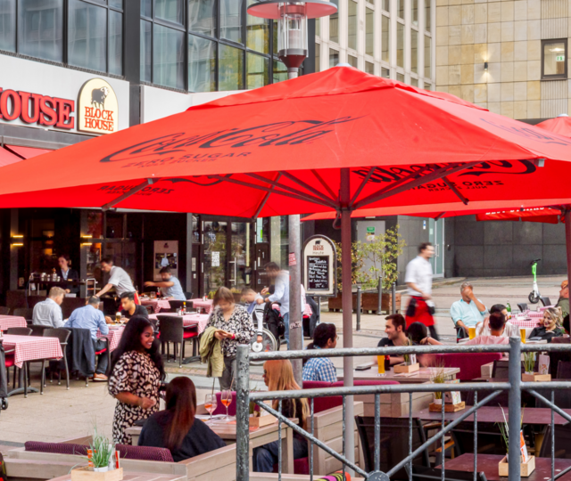 Außenansicht des BLOCK HOUSE Essen mit großer Terrasse, roten Sonnenschirmen und Gästen im Außenbereich.