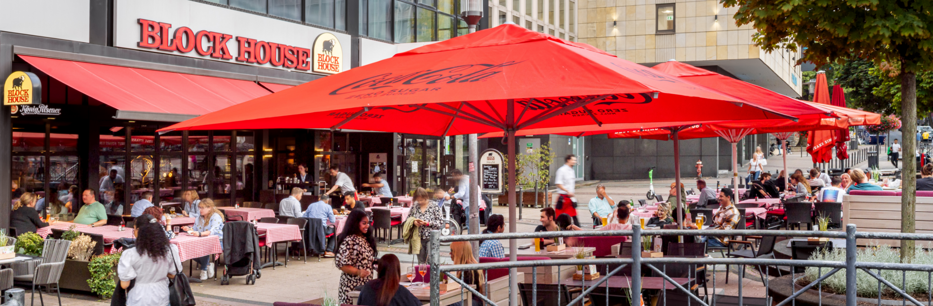 BLOCK HOUSE Essen Außenansicht des BLOCK HOUSE Essen mit großer Terrasse, roten Sonnenschirmen und Gästen im Außenbereich.
