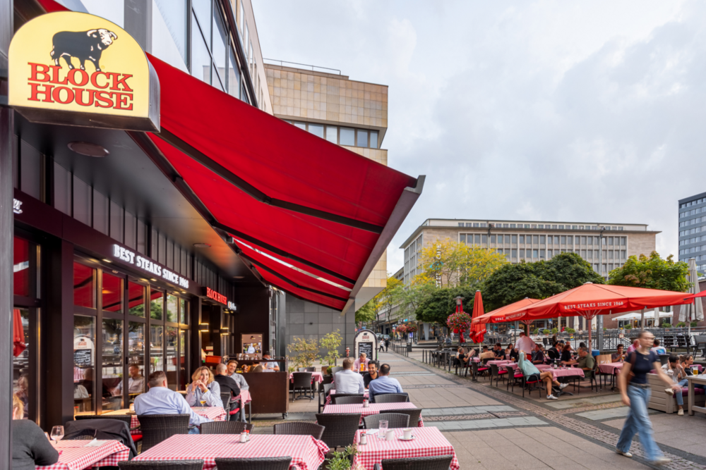 BLOCK HOUSE Essen Außenbereich des BLOCK HOUSE Essen mit Terrasse, roten Markisen und Gästen auf dem Platz.