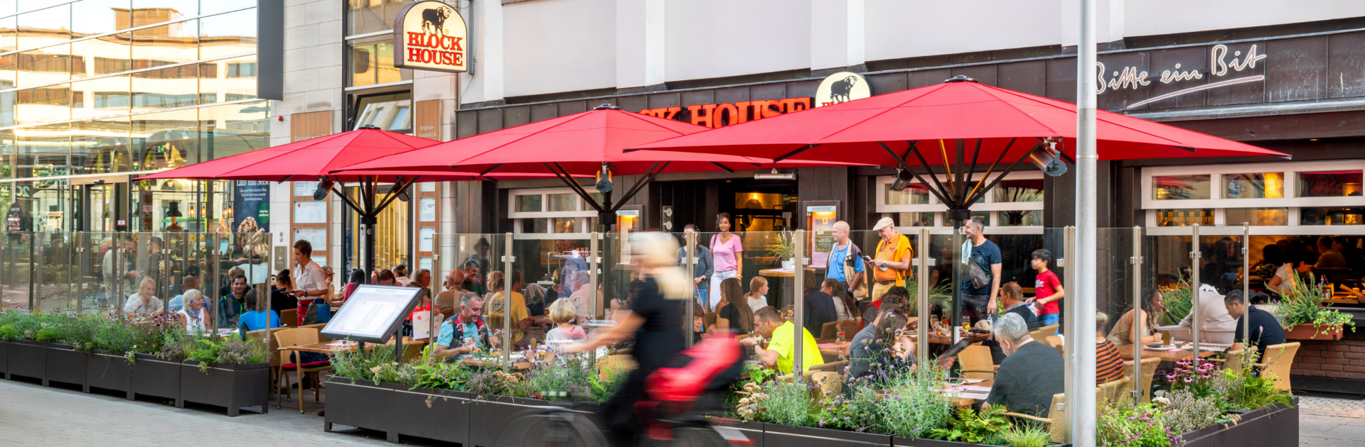 Terrasse des BLOCK HOUSE Hannover Am Kröpcke mit Gästen unter roten Sonnenschirmen in der Fußgängerzone der Innenstadt