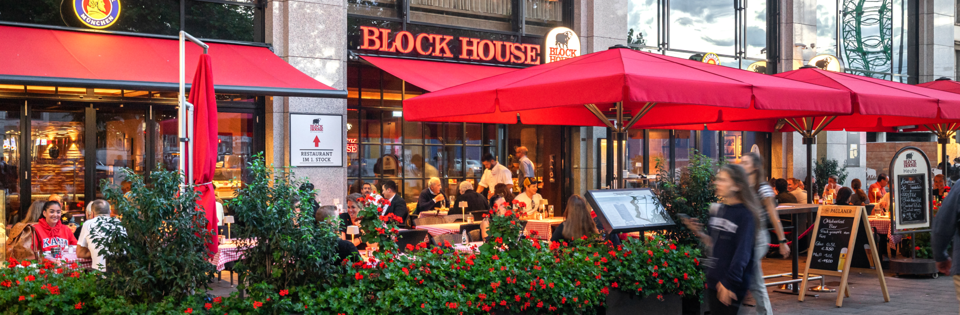 Außenansicht des BLOCK HOUSE München Elisenhof mit Terrasse, roten Sonnenschirmen und Gästen im Außenbereich.