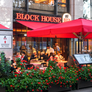 BLOCK HOUSE München Elisenhof Außenansicht des BLOCK HOUSE München Elisenhof mit Terrasse, roten Sonnenschirmen und Gästen im Außenbereich.