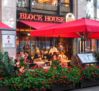 Außenansicht des BLOCK HOUSE München Elisenhof mit Terrasse, roten Sonnenschirmen und Gästen im Außenbereich.
