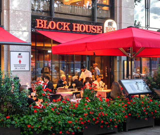 Außenansicht des BLOCK HOUSE München Elisenhof mit Terrasse, roten Sonnenschirmen und Gästen im Außenbereich.