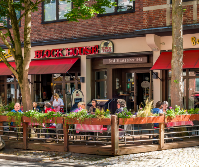 BLOCK HOUSE Restaurant in Kiel Außenansicht des BLOCK HOUSE Kiel mit Terrasse, roten Markisen und Gästen vor dem Restaurant.