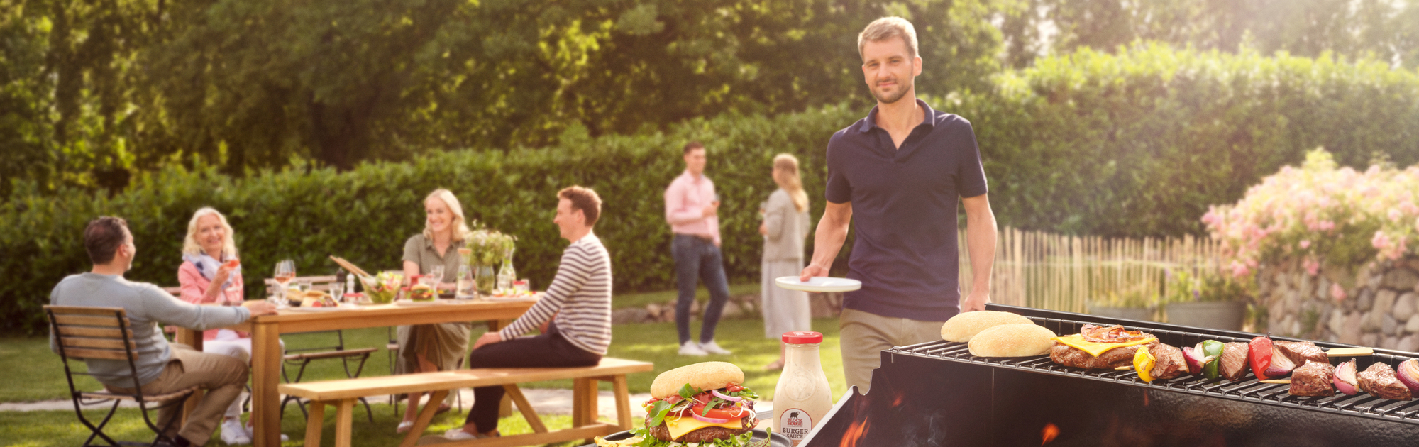 Im Vordergrund ein Grill mit BLOCK HOUSE Burgern und Sauce; links sitzen mehrere Personen an einem Tisch, im Hintergrund unterhalten sich ein Mann und eine Frau; ein weiterer Mann kommt mit seinem Teller auf den Grill zu
