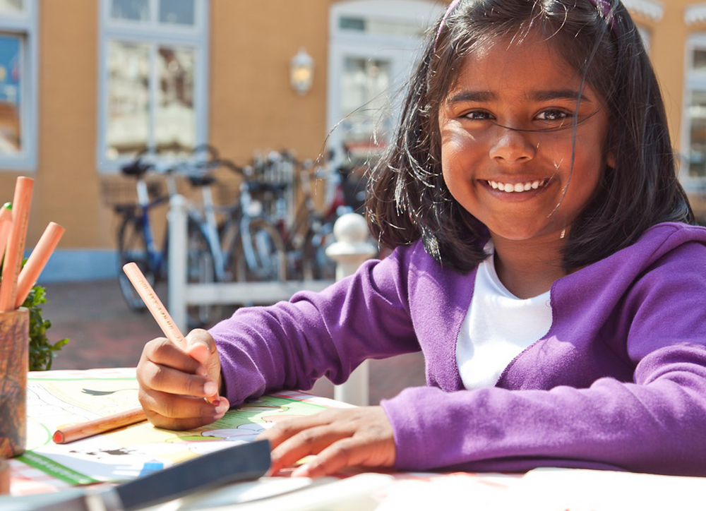 Ein Mädchen sitzt an einem Tisch im BLOCK HOUSE Restaurant. Sie lächelt in die Kamera und hält einen Buntstift in der Hand. Vor ihr liegen BLOCK HOUSE Malunterlagen. 