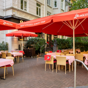 Eingangsbereich und Terrasse des BLOCK HOUSE Hamburg Winterhude mit Sonnenschirmen, Sitzplätzen und altem Baumbestand