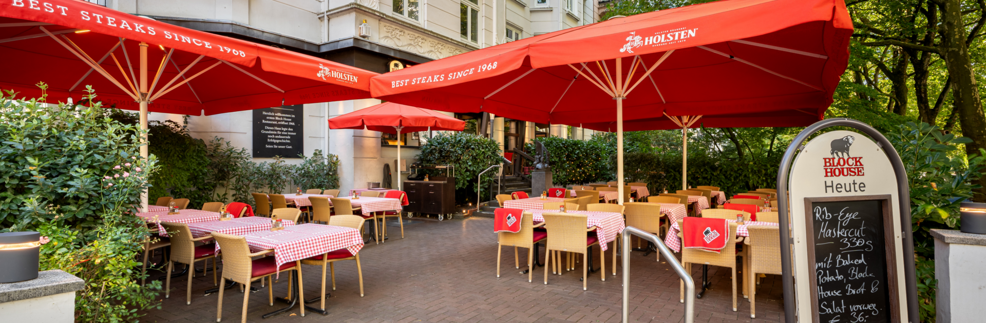 Eingangsbereich und Terrasse des BLOCK HOUSE Hamburg Winterhude mit Sonnenschirmen, Sitzplätzen und altem Baumbestand