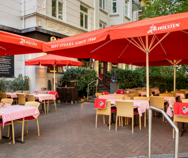 BLOCK HOUSE Hamburg Winterhude – Erstes Steakhouse seit 1968 mit Terrasse Eingang und Terrasse des BLOCK HOUSE Hamburg Winterhude mit roten Sonnenschirmen und Sitzplätzen im Grünen.