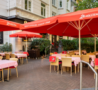 BLOCK HOUSE Hamburg Winterhude – Erstes Steakhouse seit 1968 mit Terrasse Eingang und Terrasse des BLOCK HOUSE Hamburg Winterhude mit roten Sonnenschirmen und Sitzplätzen im Grünen.